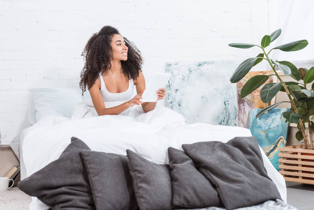 vrolijke Afro-Amerikaanse jonge vrouw met behulp van digitale tablet in bed tijdens ochtend tijd thuis - Foto, afbeelding