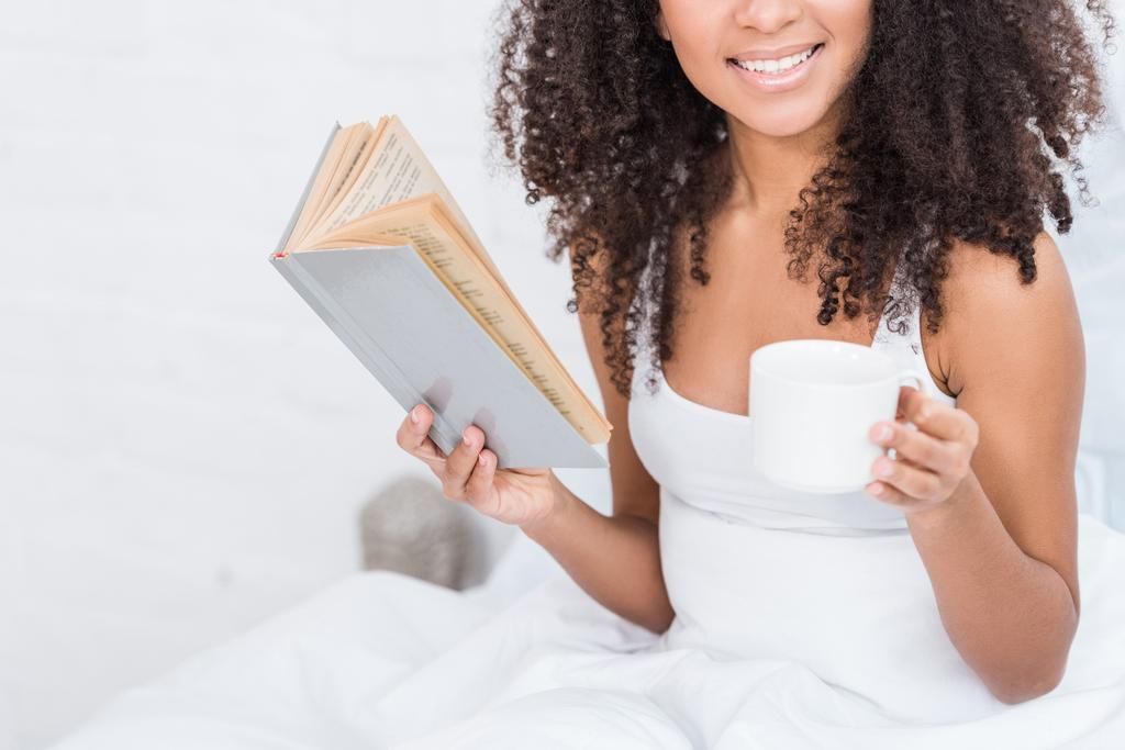 vista parziale della ragazza afro-americana che tiene una tazza di caffè e legge un libro a letto durante il mattino a casa
 - Foto, immagini