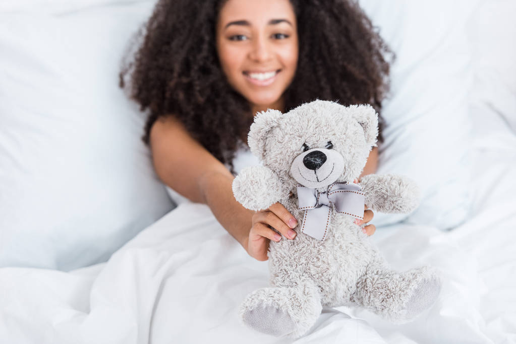selective focus of african american girl laying in bed and showing teddy bear at home - Photo, Image