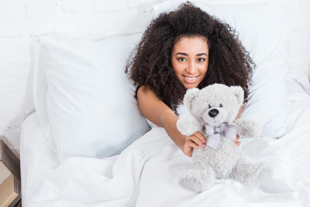 μεγάλη γωνία προβολής του νεαρή αφρικανική αμερικανική γυναίκα βάζοντας στο κρεβάτι και δείχνει το αρκουδάκι στο σπίτι - Φωτογραφία, εικόνα