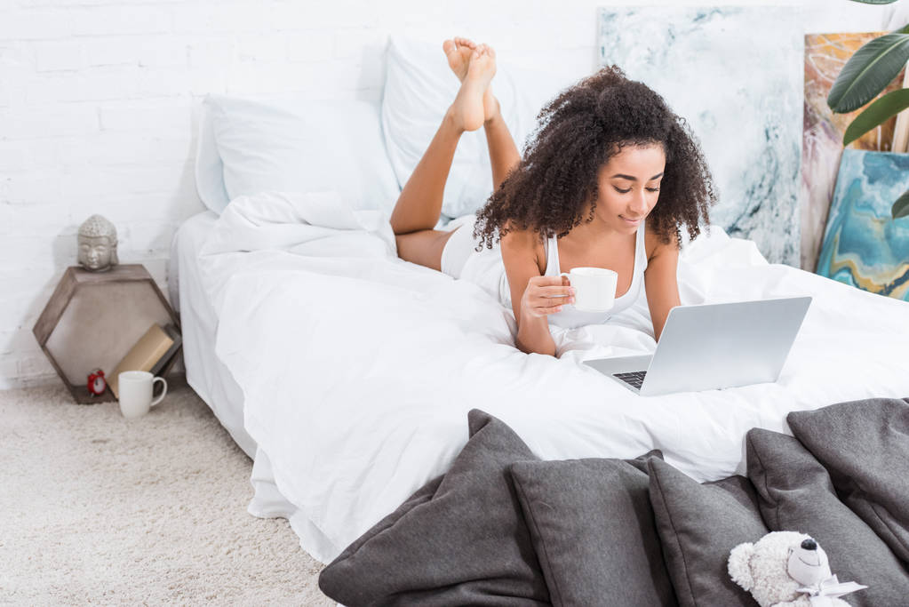 afrikanisch-amerikanisches lockiges Mädchen mit Kaffeetasse und Laptop im Bett während der Morgenzeit zu Hause - Foto, Bild