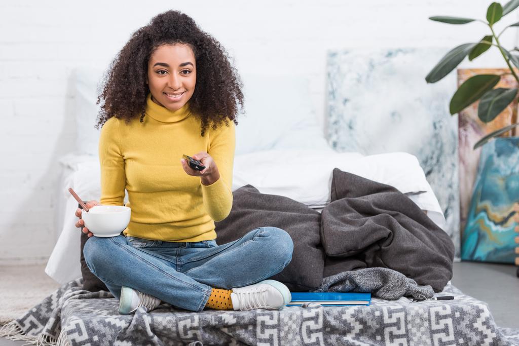 pretty stylish african american girl holding bowl with food and watching tv at home - Photo, Image