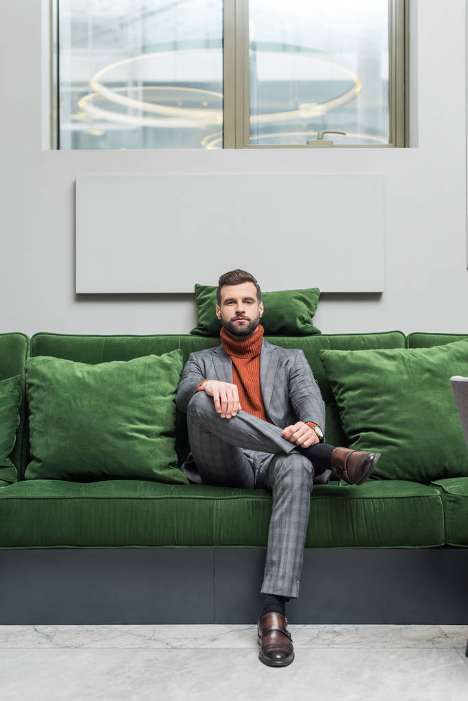man in formal wear with legs crossed sitting on green sofa and looking at camera - Photo, Image