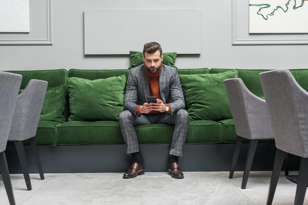 ernster gutaussehender Mann in formeller Kleidung sitzt auf grünem Sofa und benutzt Smartphone - Foto, Bild