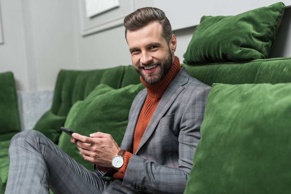 χαμογελώντας όμορφος άνδρας στην επίσημη ένδυση κάθεται στο πράσινο καναπέ και χρησιμοποιώντας το smartphone - Φωτογραφία, εικόνα
