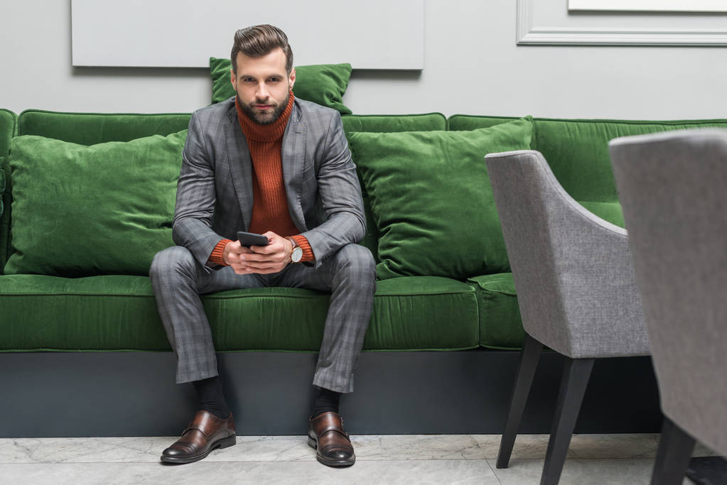 hombre guapo en ropa formal sentado en un sofá verde, mirando a la cámara y utilizando el teléfono inteligente
 - Foto, imagen