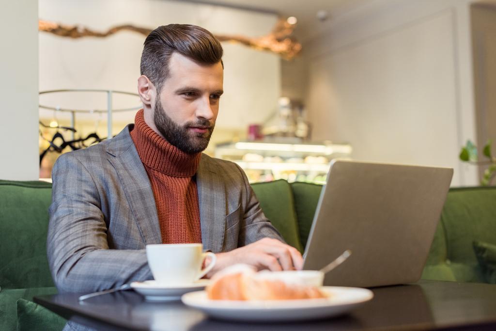 focused handsome businessman in formal wear sitting at table and typing on laptop in restaurant - Photo, Image