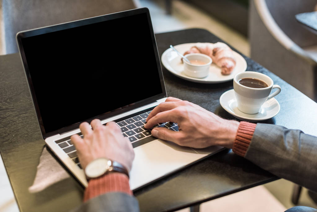 bijgesneden weergave van zakenman zittend aan tafel met koffie en croissant tijdens het typen op laptop met leeg scherm in restaurant - Foto, afbeelding