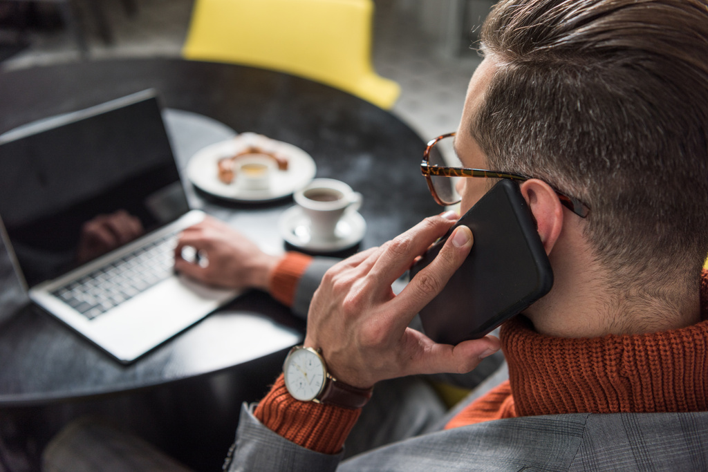 close up de empresário bonito em óculos e desgaste formal sentado à mesa, falando no smartphone e digitando no laptop no restaurante
 - Foto, Imagem