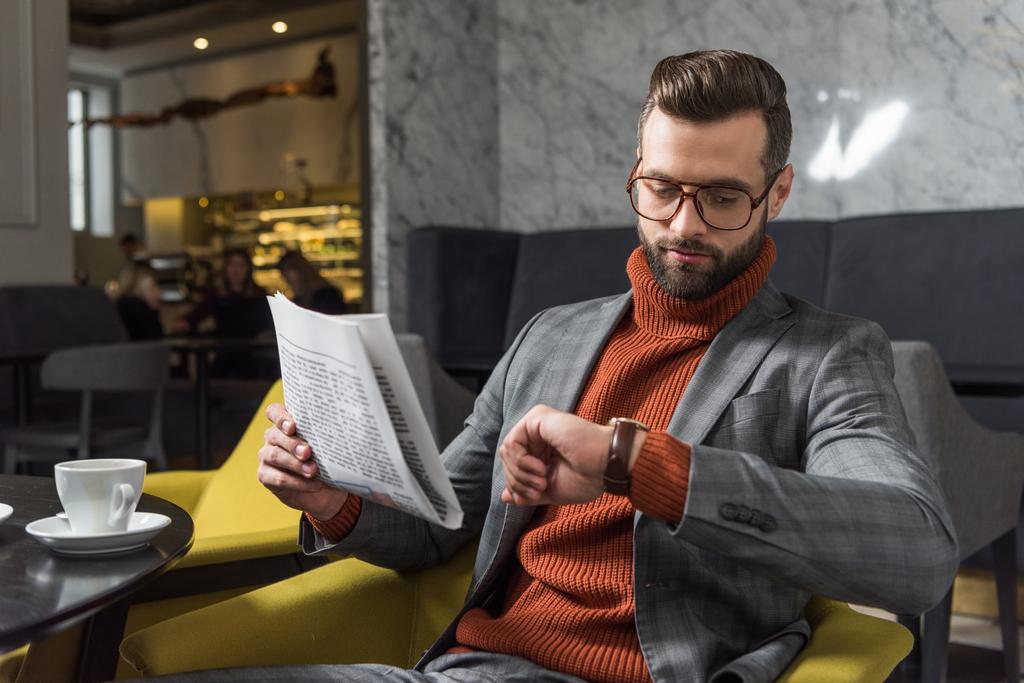 stylish man in formal wear and glasses holding newspaper and looking at watch in restaurant - Photo, Image