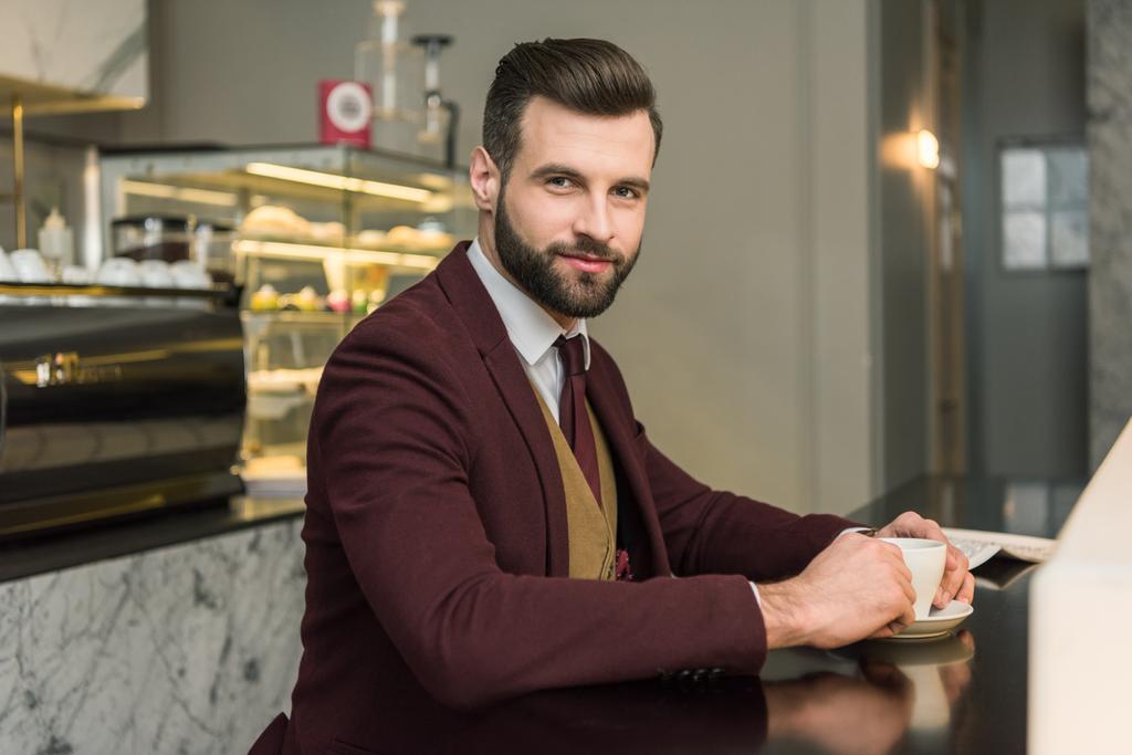schöner Geschäftsmann in formeller Kleidung, der in die Kamera schaut und mit Kaffee im Restaurant am Tisch sitzt - Foto, Bild