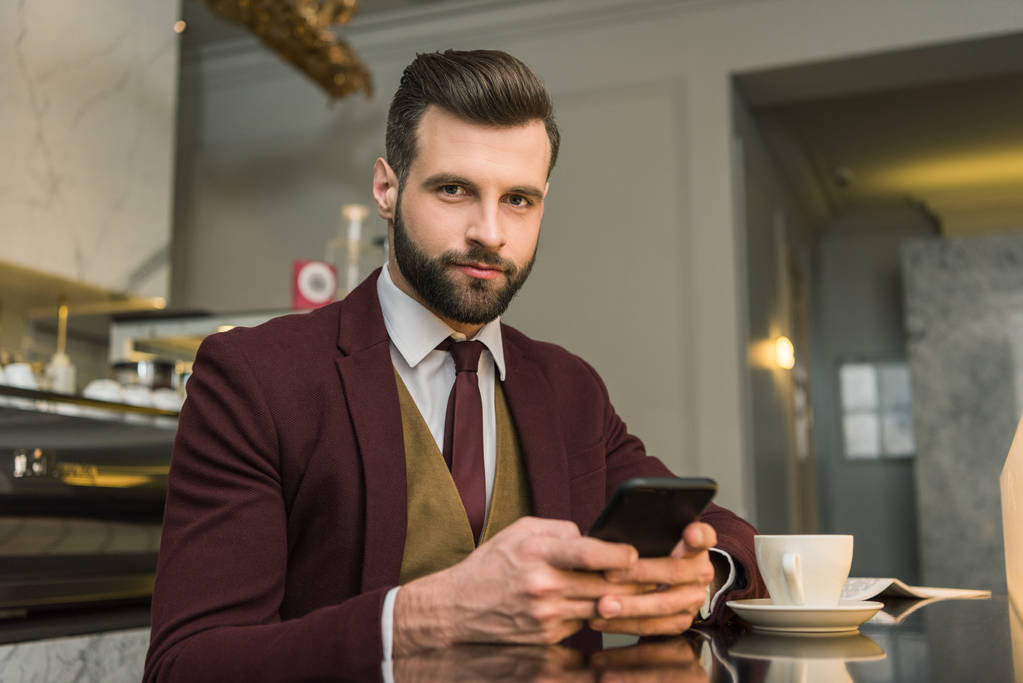 vážně hezký podnikatel v oblasti formální oblečení sedí u stolu s kávou a pomocí smartphone v restauraci - Fotografie, Obrázek