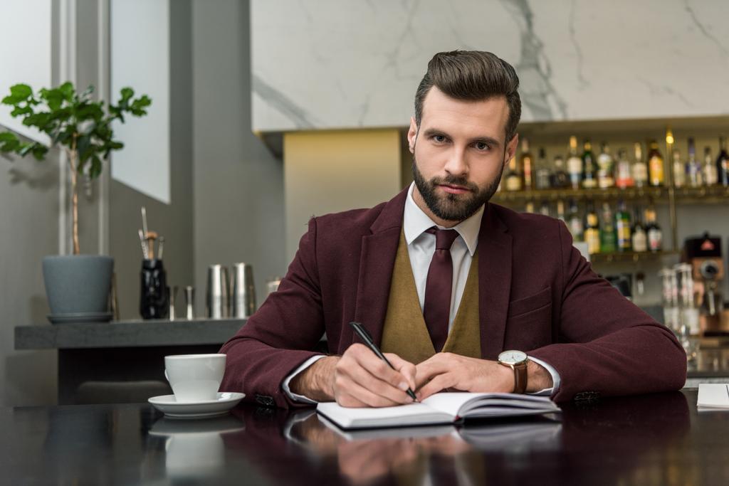 serious businessman in formal wear sitting and writing in notebook at table in restaurant - Photo, Image