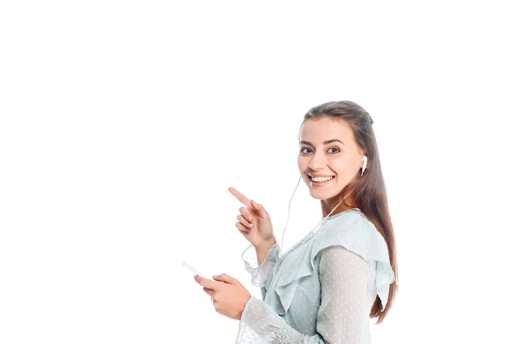 side view of smiling woman with smartphone pointing away while listening music in earphones isolated on white - Photo, Image