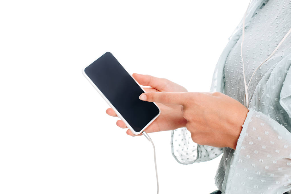 gedeeltelijke weergave van woman wijzend op smartphone terwijl het luisteren muziek in oortelefoons geïsoleerd op wit - Foto, afbeelding