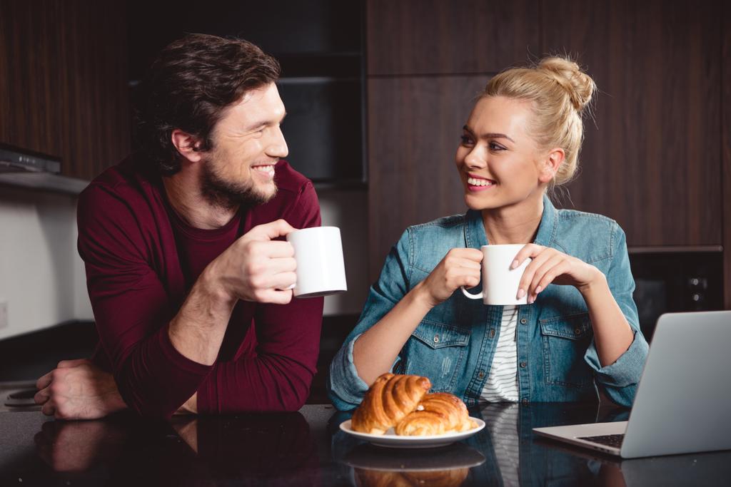 happy couple holding coffee cups and looking at each other in kitchen - Photo, Image