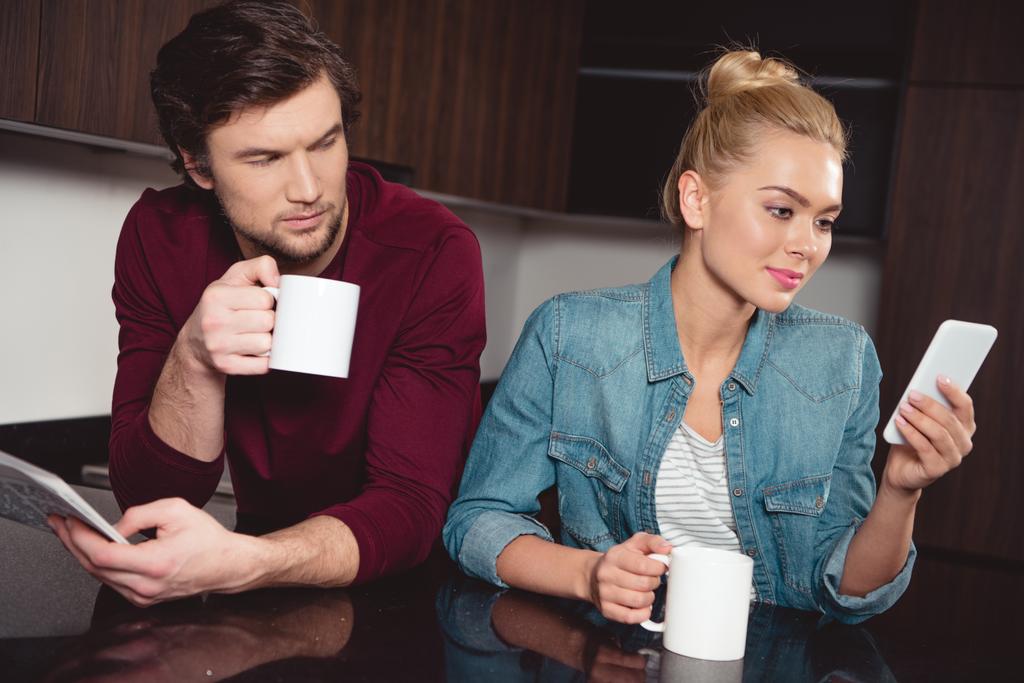jealous husband holding coffee cup and looking at wife using smartphone in kitchen - Photo, Image