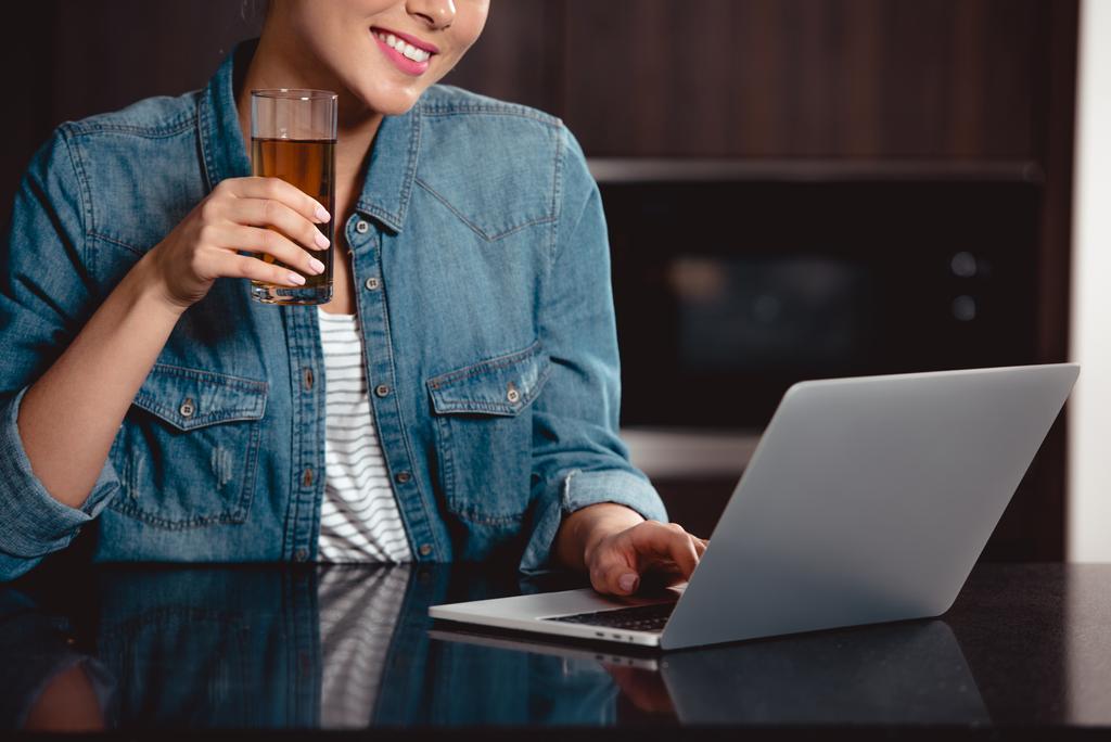 обрізаний вид усміхненої дівчини, що тримає склянку соку і друкує на клавіатурі ноутбука вдома
 - Фото, зображення