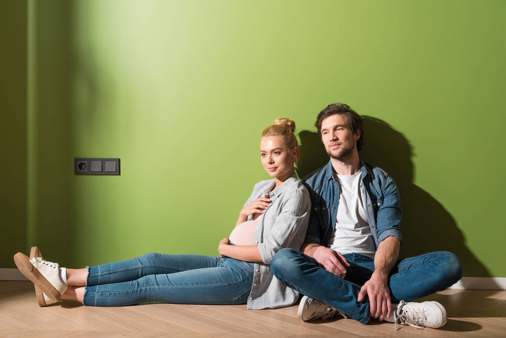 задумлива вагітна дівчина сидить на підлозі з чоловіком на зеленій стіні
 - Фото, зображення