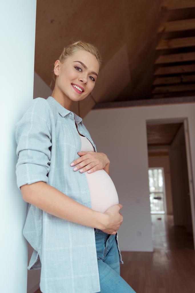 Beautiful pregnant girl touching belly with both hands and looking at camera in apartment - Photo, Image