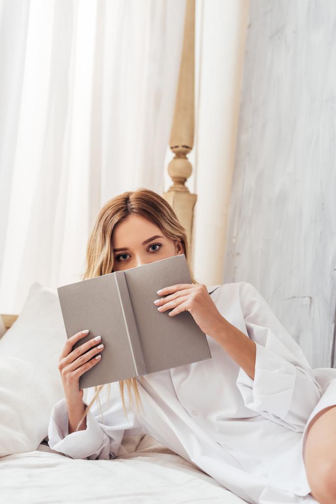 Blondynka, młoda kobieta, obejmujących twarz przez książki i patrząc na kamery w łóżku w domu - Zdjęcie, obraz