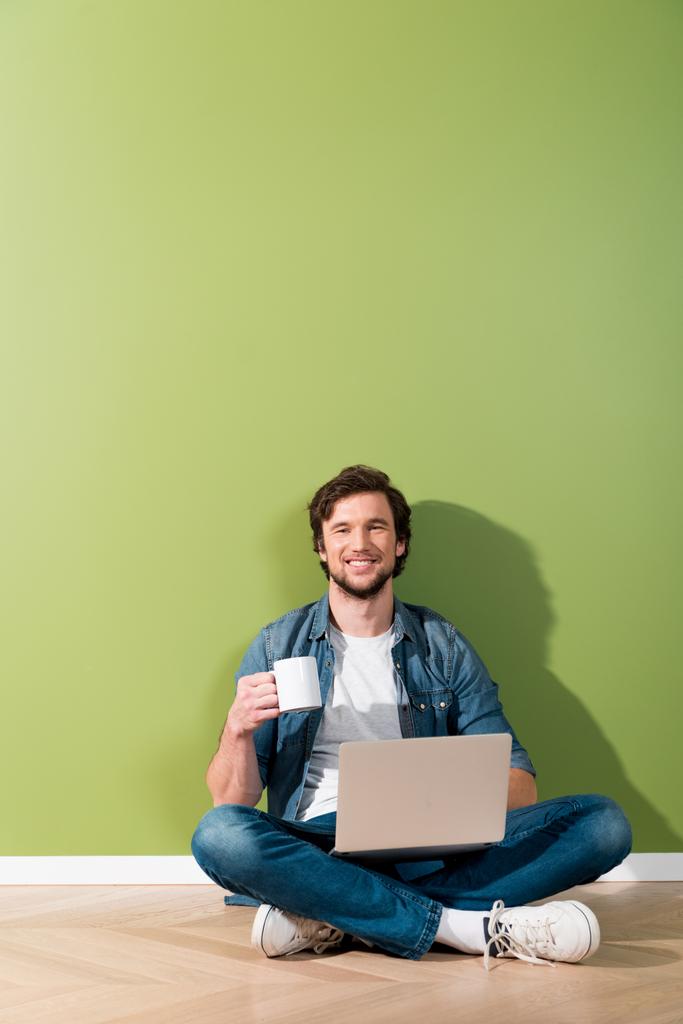 homme souriant tenant tasse de café et ordinateur portable et assis sur le sol et regardant la caméra
 - Photo, image