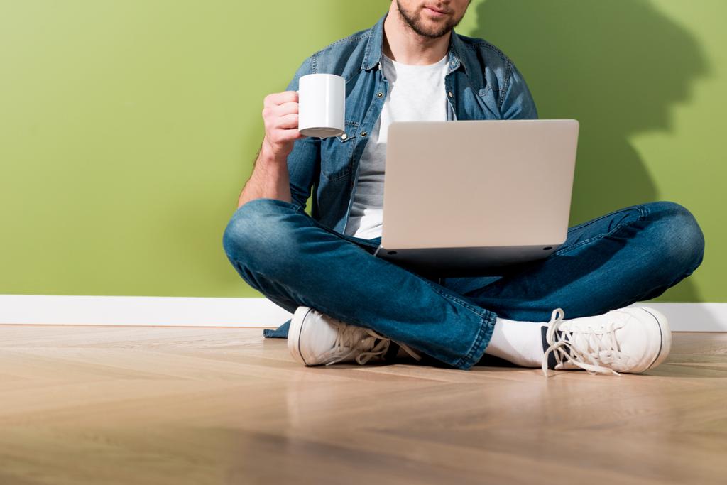 cropped view of man holding coffee cup and laptop and sitting on floor by green wall - Photo, Image