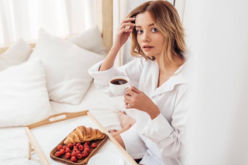 selectieve aandacht van jonge vrouw houden van koffiekopje terwijl zittend op bed met ontbijt op dienblad - Foto, afbeelding