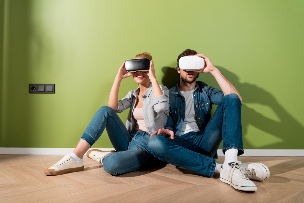 έκπληκτος κορίτσι και ο άνθρωπος κάθεται στο πάτωμα και κρατώντας τις κάσκες εικονικής πραγματικότητας στο κεφάλι  - Φωτογραφία, εικόνα