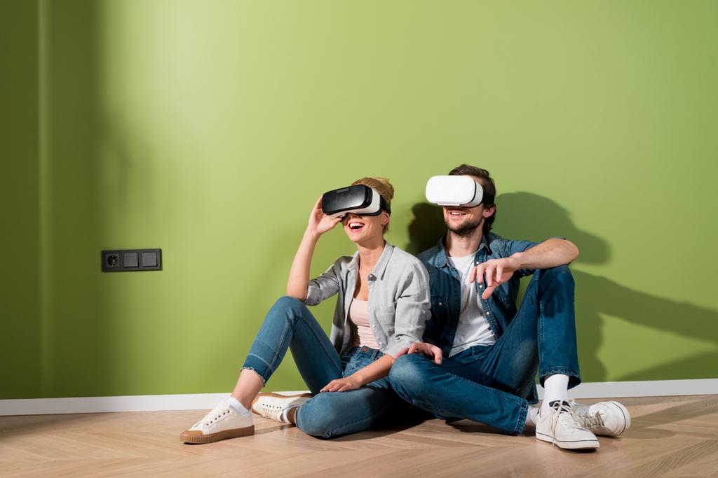 удивленная пара с гарнитурами виртуальной реальности на головах, сидящих на полу за зеленой стеной
 - Фото, изображение