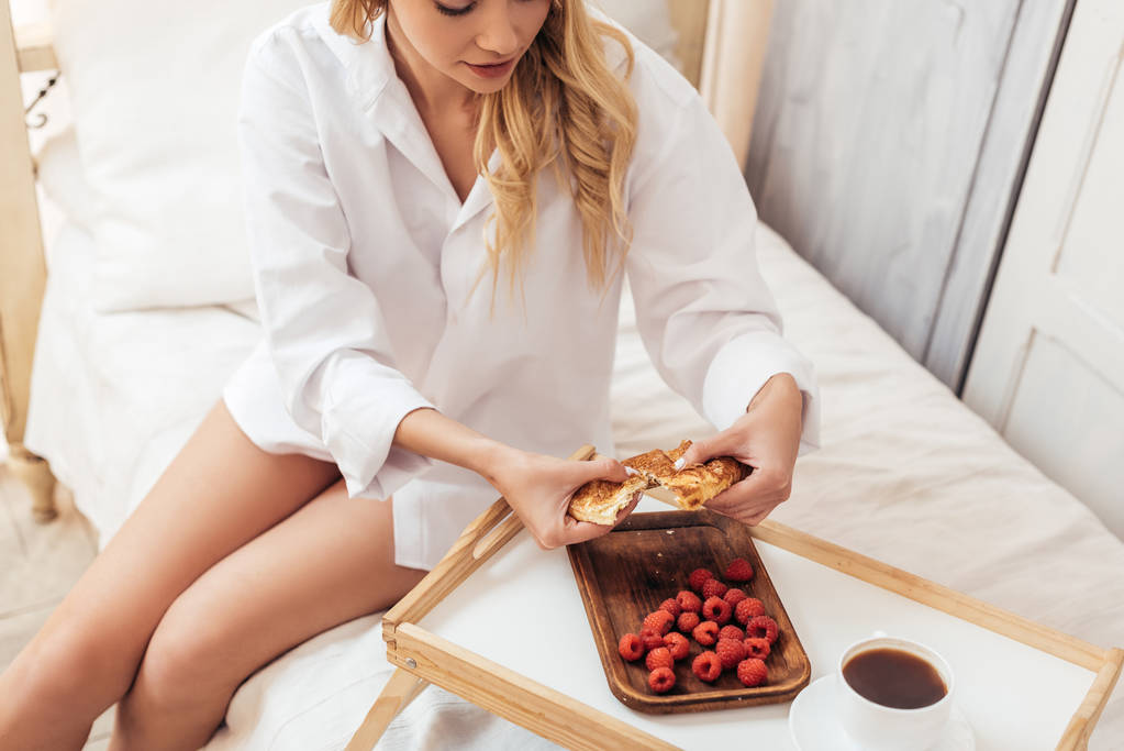 gedeeltelijke weergave van vrouw scheuren croissant in bed met ontbijt op dienblad tijdens ochtend tijd thuis - Foto, afbeelding