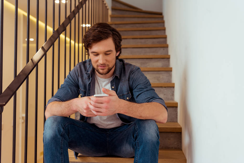 pohledný muž sedí na schodech a použití smartphone doma - Fotografie, Obrázek