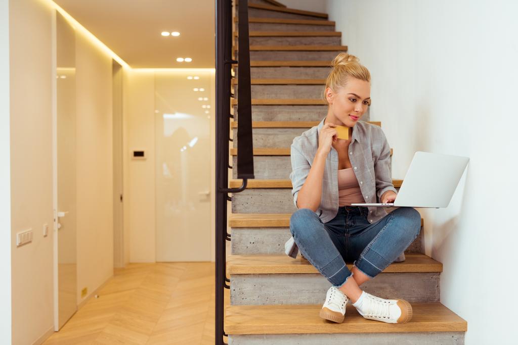 attractive girl sitting on stairs, holding credit card and using laptop - Photo, Image