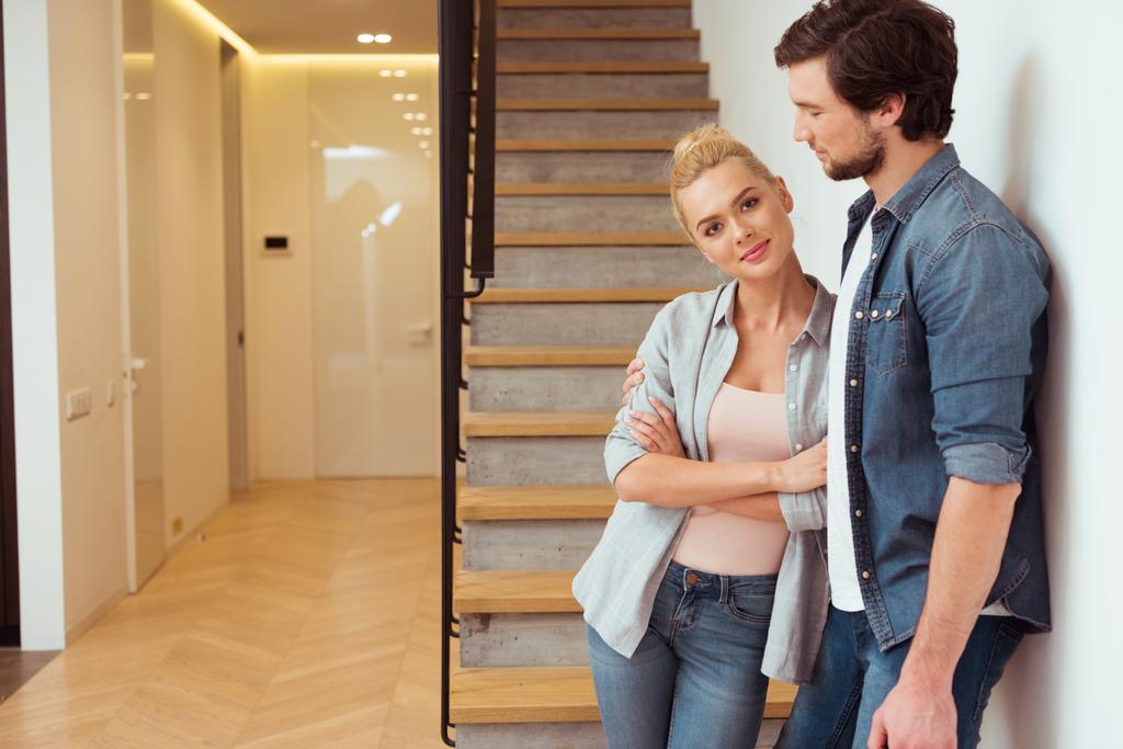 attractive girl looking at camera and standing with husband near stairs at home - Photo, Image