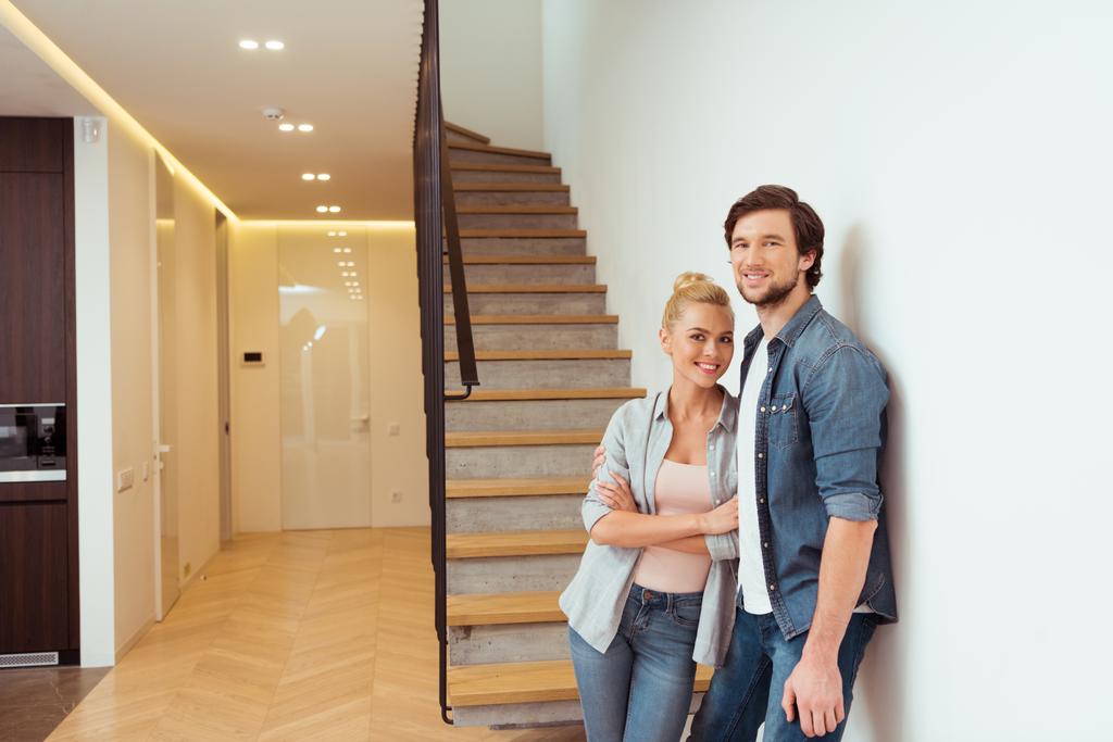 alegre pareja de pie cerca de escaleras y mirando a la cámara en casa
 - Foto, Imagen