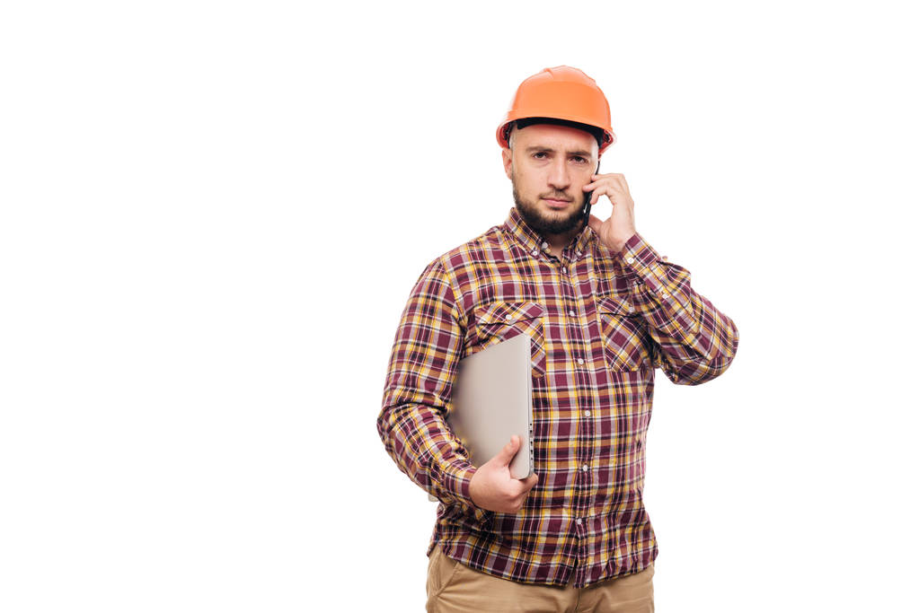 Glücklicher Bauarbeiter mit orangefarbenem Schutzhelm, der einen Laptop in der Hand hält und auf weißem Hintergrund isoliert telefoniert. Kopierraum für Text. Zeit zum Arbeiten.  - Foto, Bild