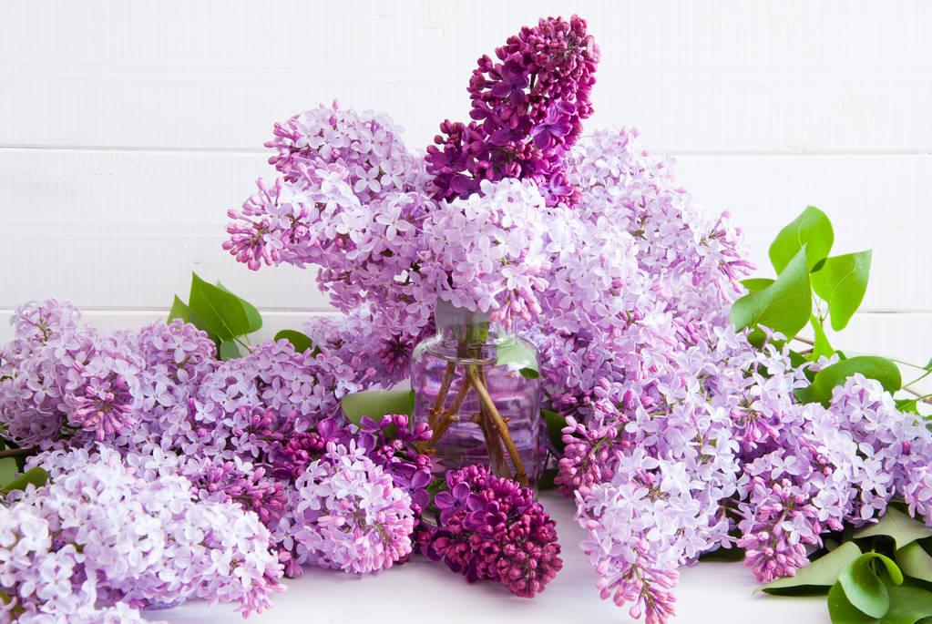 Violetti kukkia lila lehtiä lasipullossa valkoisella puupohjalla
 - Valokuva, kuva