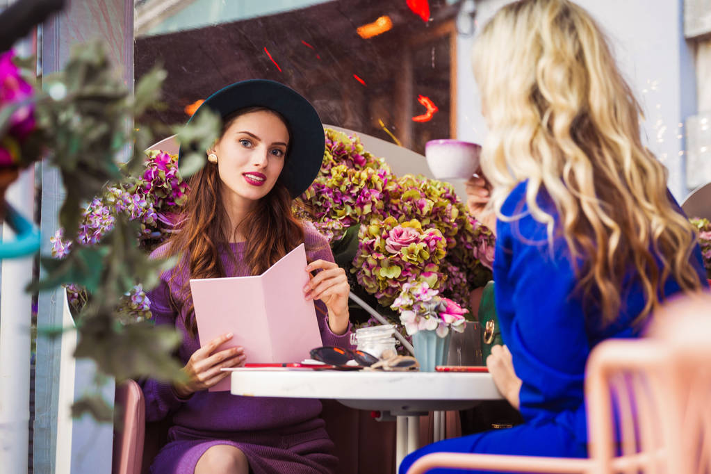 Dvě elegantní mladé ženy sedí ve venkovní restauraci a jeden pití šálku čaje a další Svobodník v menu - Fotografie, Obrázek