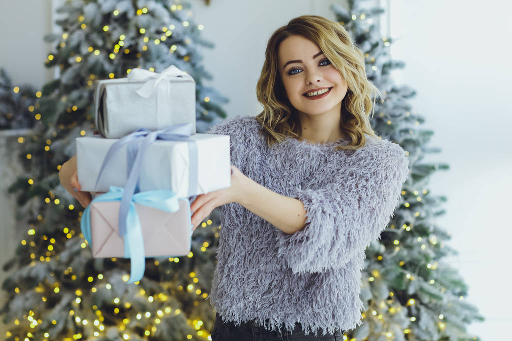 Jeune belle femme blonde dans un pull gris avec un cadeau sur les mains près du Nouvel An, arbre de Noël avec des cadeaux
 - Photo, image