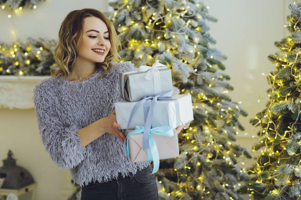 Giovane bella donna bionda in un maglione grigio con un regalo sulle mani vicino al nuovo anno, albero di Natale con regali
 - Foto, immagini
