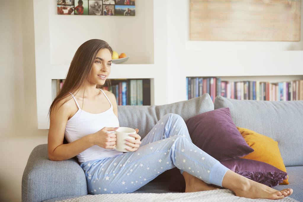Aufnahme der attraktiven jungen Frau im Schlafanzug, die ihren Morgentee trinkt, während sie es sich auf dem Sofa in ihrer Wohnung gemütlich macht.  - Foto, Bild