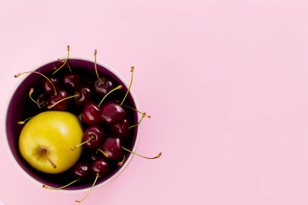 Světlé zralé třešně a žluté šťavnaté jablko v talíř na pastelově růžové pozadí. - Fotografie, Obrázek