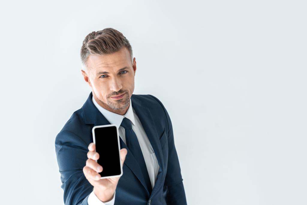 Высокоугольный вид красивого бизнесмена, показывающего смартфон с затемненным экраном и смотрящего на камеру изолированную на белом
 - Фото, изображение