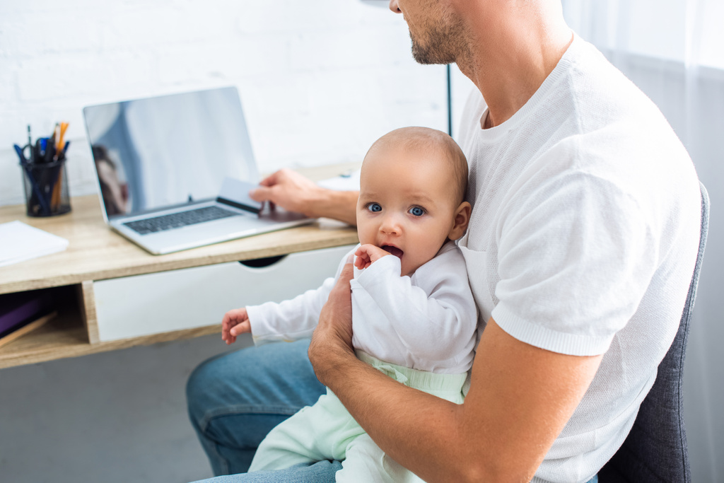 Vater sitzt am Computertisch mit Kreditkarte für Online-Einkäufe und hält entzückende kleine Tochter zu Hause - Foto, Bild