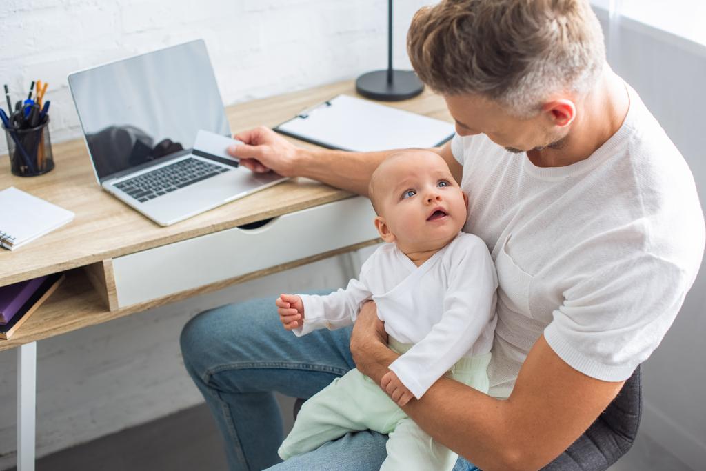 father sitting at computer desk with credit card for online shopping and holding adorable baby daughter at home - Photo, Image