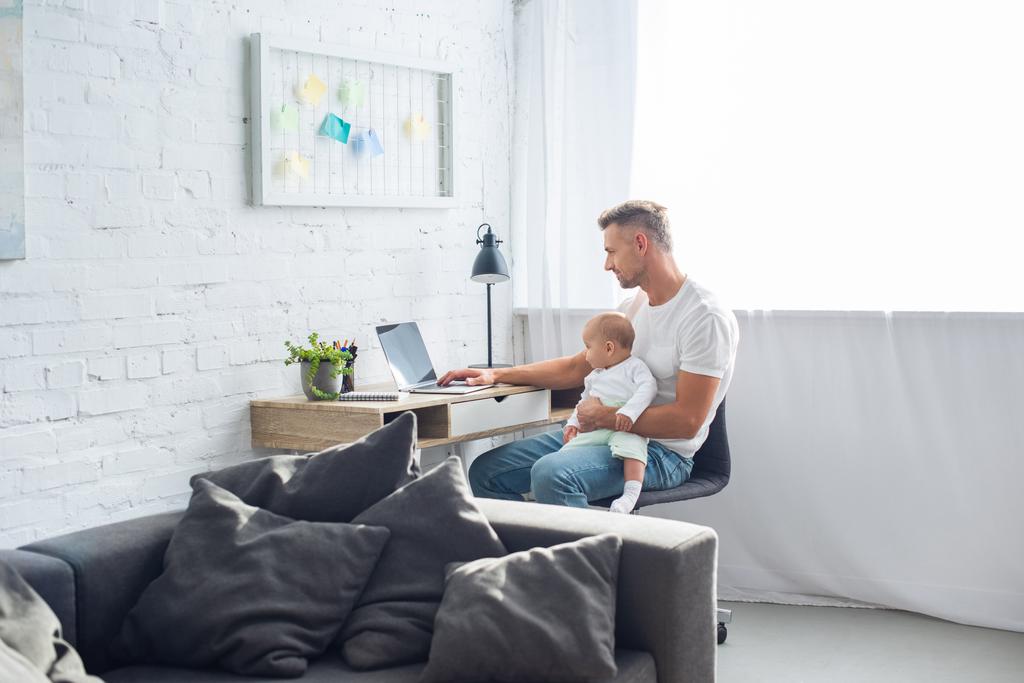 father sitting at desk, using laptop and holding baby daughter in modern apartment - Photo, Image