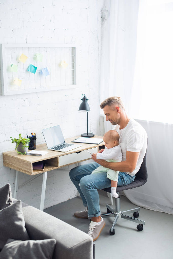 père assis au bureau de l'ordinateur, à l'aide d'un smartphone et tenant bébé fille dans le salon
 - Photo, image