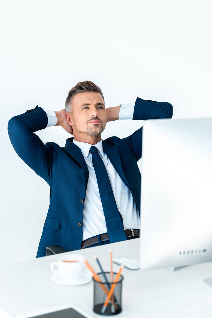 красивый бизнесмен в костюме сидит за столом с компьютером и держит руки за головой изолированы на белом
 - Фото, изображение
