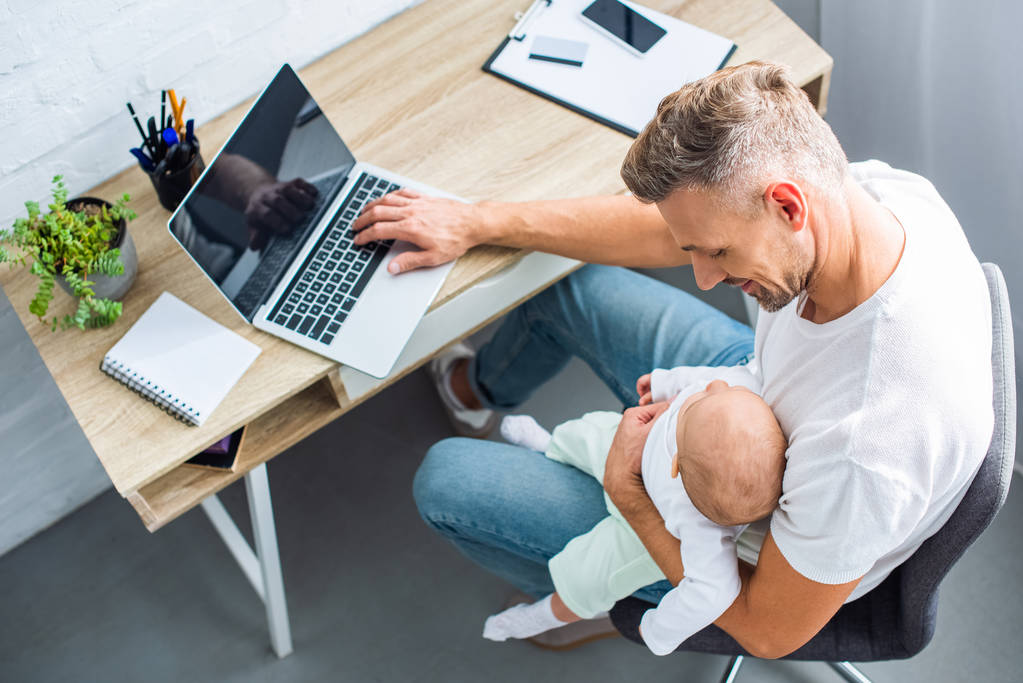 father sitting at desk and using laptop with blank screen while holding baby daughter at home - Photo, Image