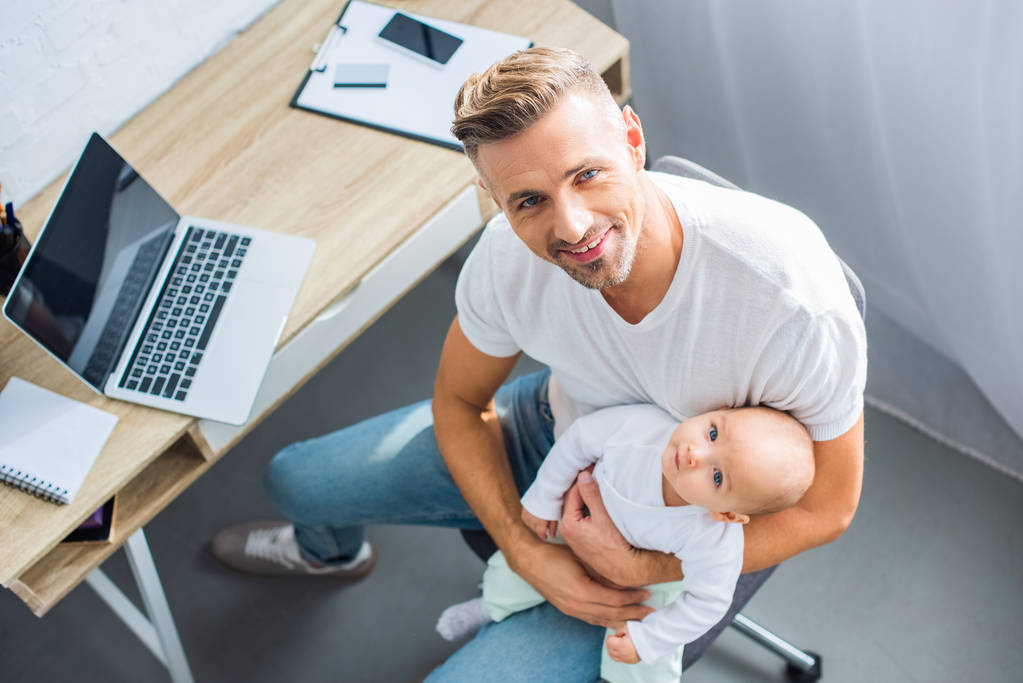 father sitting at computer desk, holding baby daughter and looking at camera at home - Photo, Image
